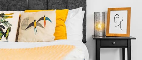 A neatly made bed with a striped blanket, a grey upholstered headboard, and decorative pillows. There is a bedside table with a framed picture and a candle.