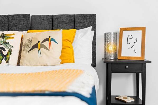 A neatly made bed with a striped blanket, a grey upholstered headboard, and decorative pillows. There is a bedside table with a framed picture and a candle.