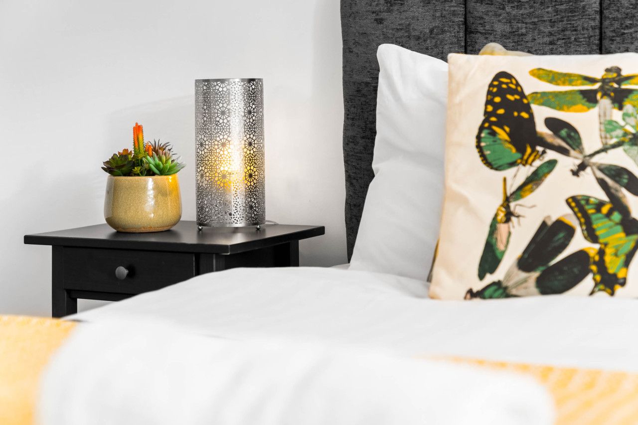 Een netjes opgemaakt bed met een wit kussen met een vlinderprint, een kleine potplant en een decoratieve wandlamp op een nachtkastje.