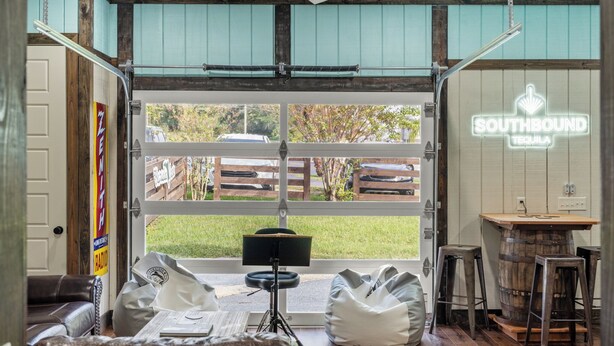 A cozy interior with a large open garage door, a bar area, and a seating area with bean bags and a leather couch. - "The Florida Cracker "THE GREEN ROOM" READ our five stars review.. (Brooksville)