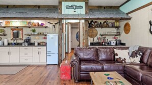 A cozy living room with a brown leather couch, a red suitcase, and a wooden coffee table. There is a kitchen area with a refrigerator, shelves with various items, and a sign that reads "Florida Cracker." - "The Florida Cracker "THE GREEN ROOM" READ our five stars review.. (Brooksville)