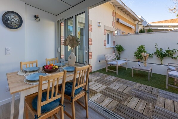 A modern outdoor dining area with a wooden table and chairs, a bowl of fruit, and a decorative vase.