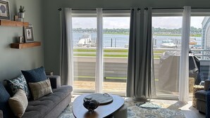 A living room with a grey sofa, a coffee table, and a view of a marina through large windows. - Wonderful 3 Bedroom Condo in Beautiful Spring Lake, On the River (Spring Lake)