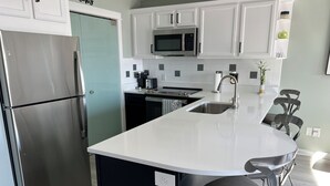 A modern kitchen with a stainless steel refrigerator, a microwave, a dishwasher, and a sink. There is a white countertop and a black island with a chair. - Wonderful 3 Bedroom Condo in Beautiful Spring Lake, On the River (Spring Lake)