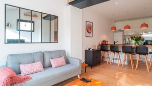 Una sala de estar moderna con un sofá gris, una mesa de centro de madera, y una cocina con taburetes altos y lámparas colgantes.