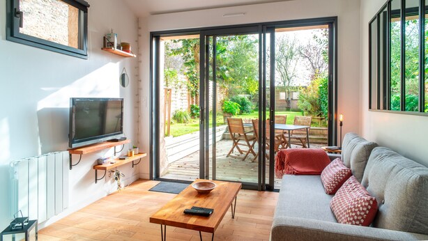 Una sala de estar moderna con un sofá, una mesa de centro de madera, un televisor montado en la pared y un ventanal corredero que da a un patio exterior con una mesa y sillas.