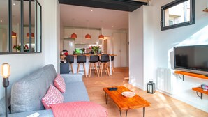 Una sala moderna con sofá, una mesa de centro de madera y televisor. Se observa una cocina con juego de comedor y lámparas colgantes.