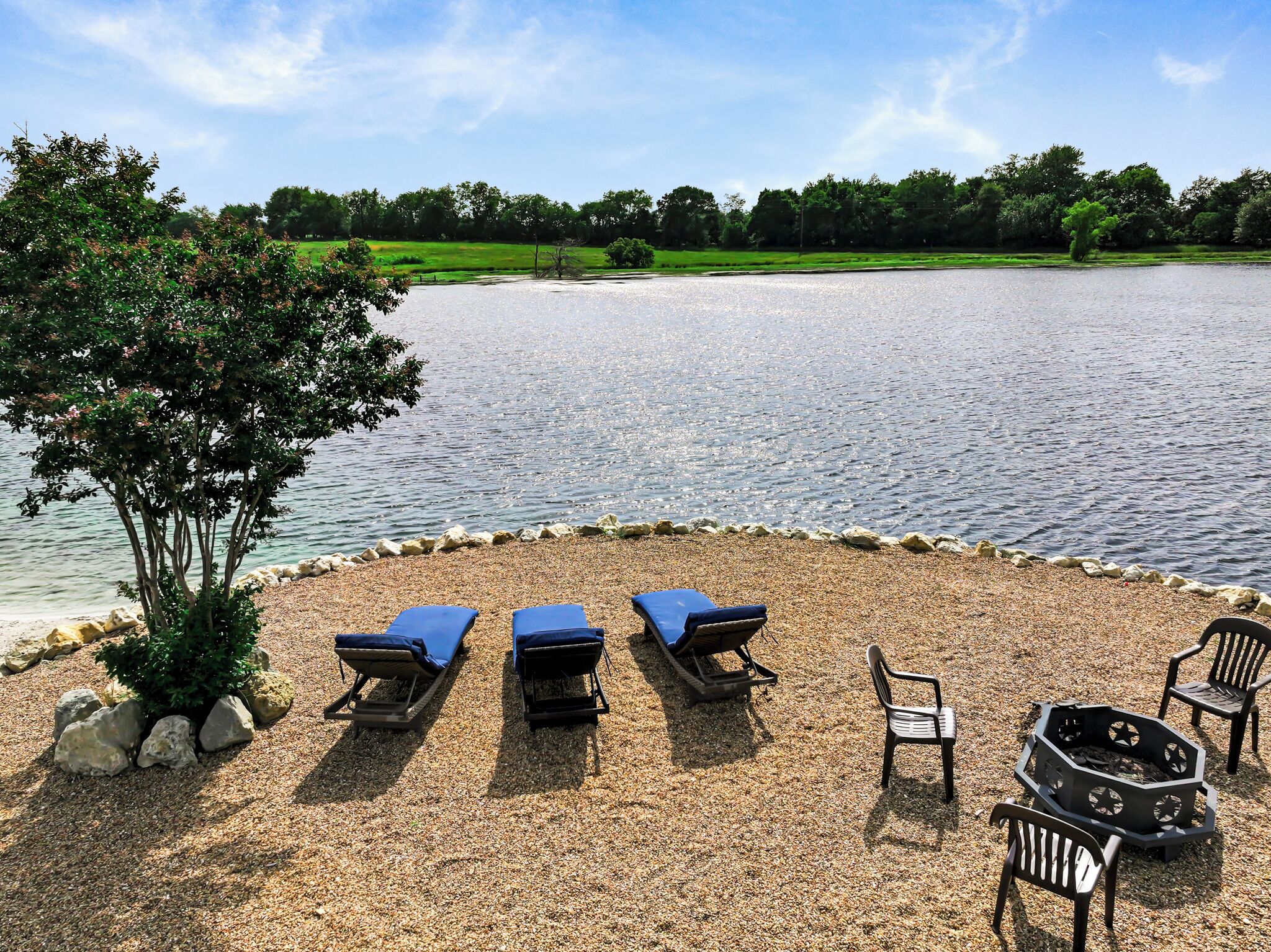 A lakeside seating area with three lounge chairs and a fire pit.