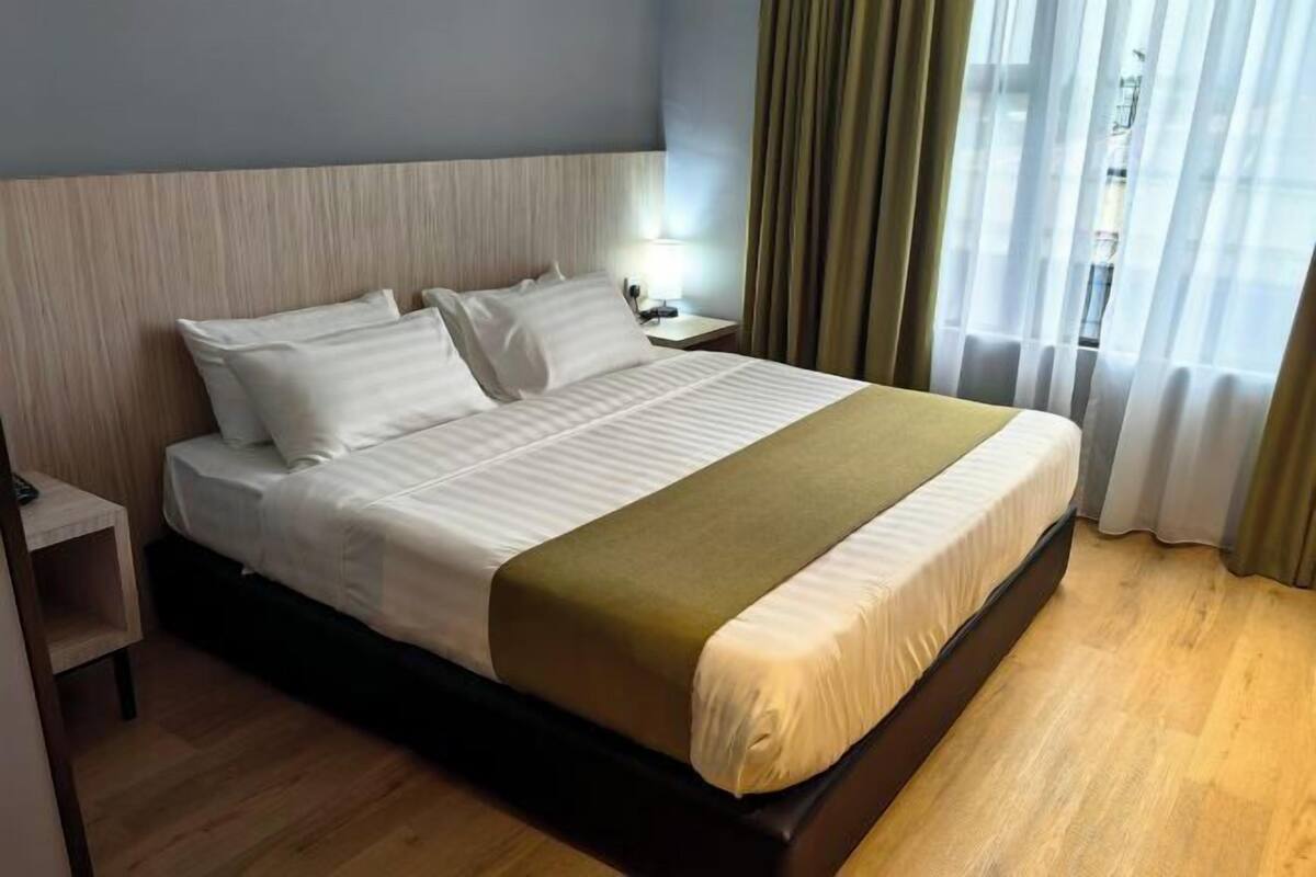 A neatly made bed with white linens and a green throw, a bedside table with a lamp, and a view of the outdoors through sheer curtains.
