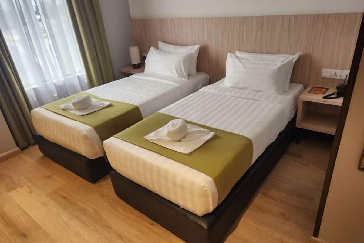 A hotel room with two beds, each with a green bedspread and white pillows. There is a nightstand with a lamp and a phone.