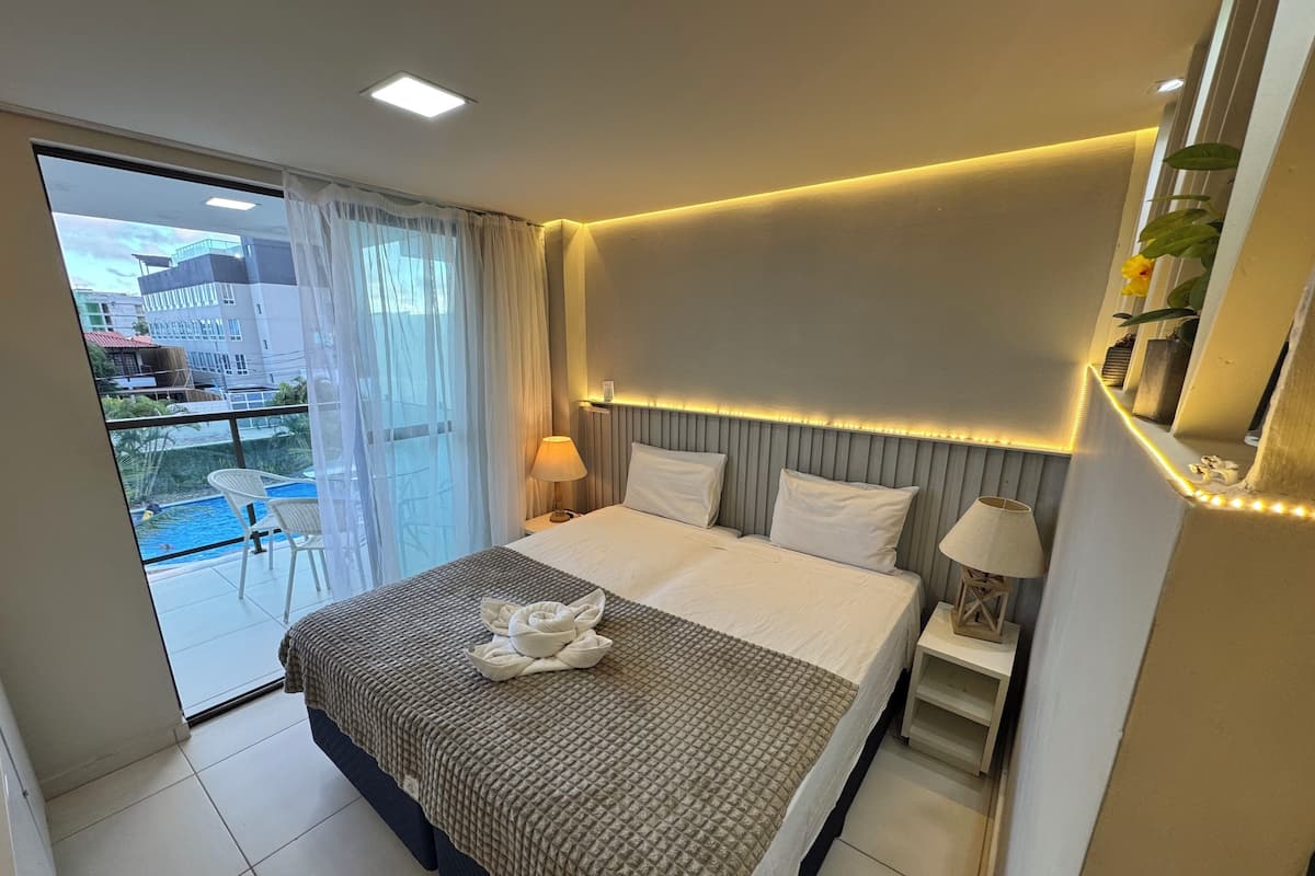 A hotel room with a bed, a bedside table with a lamp, a window with curtains, and a view of a pool and buildings outside.