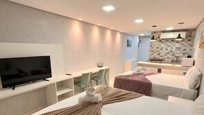 Moderni hotellihuone, jossa on suuri sänky, työpöytä, jossa on litteänäyttöinen televisio, pieni keittiö sekä oleskelualue tuoleineen.