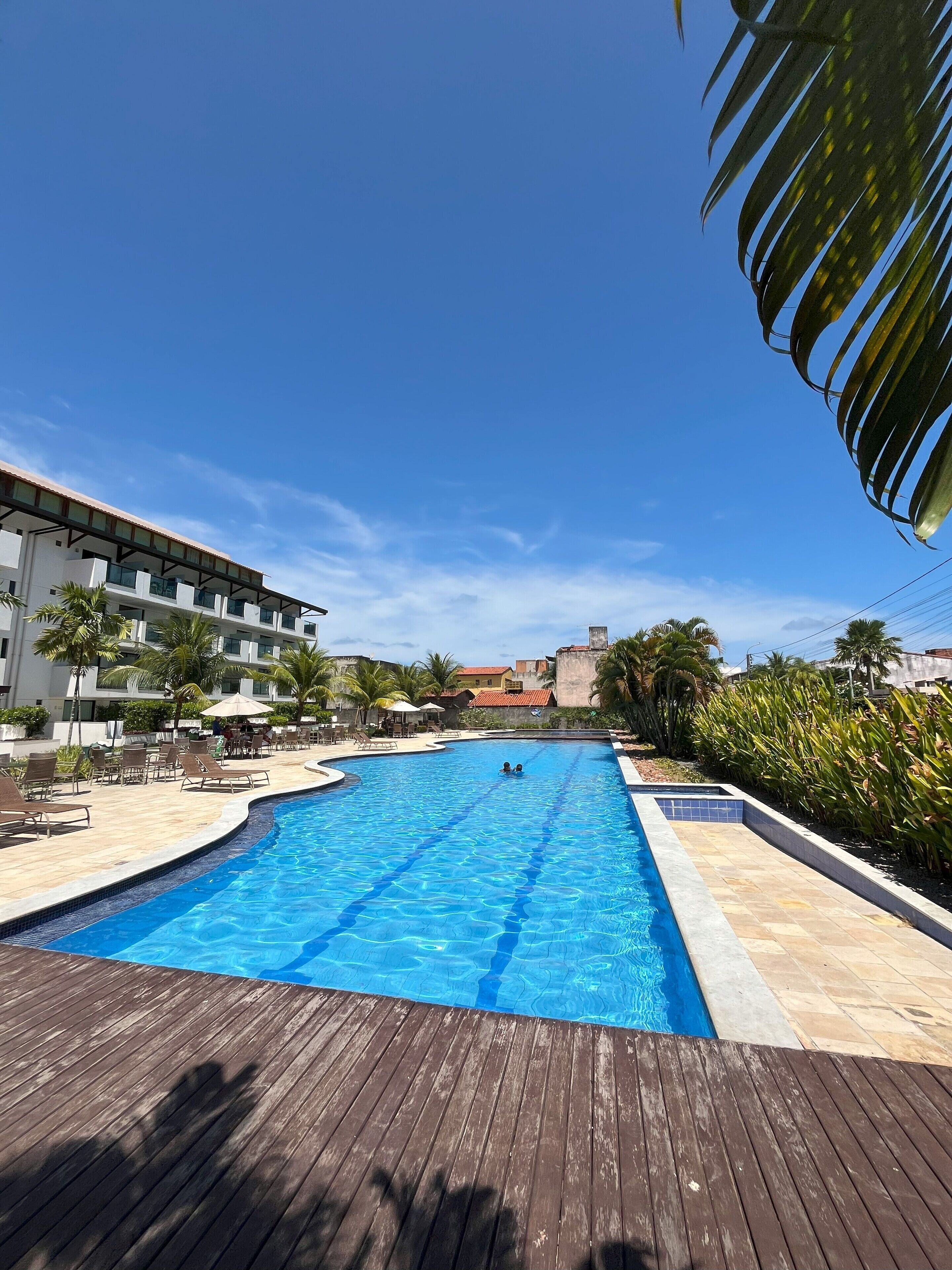 A swimming pool with a wooden deck, surrounded by palm trees and lounge chairs, with a hotel building in the background.
