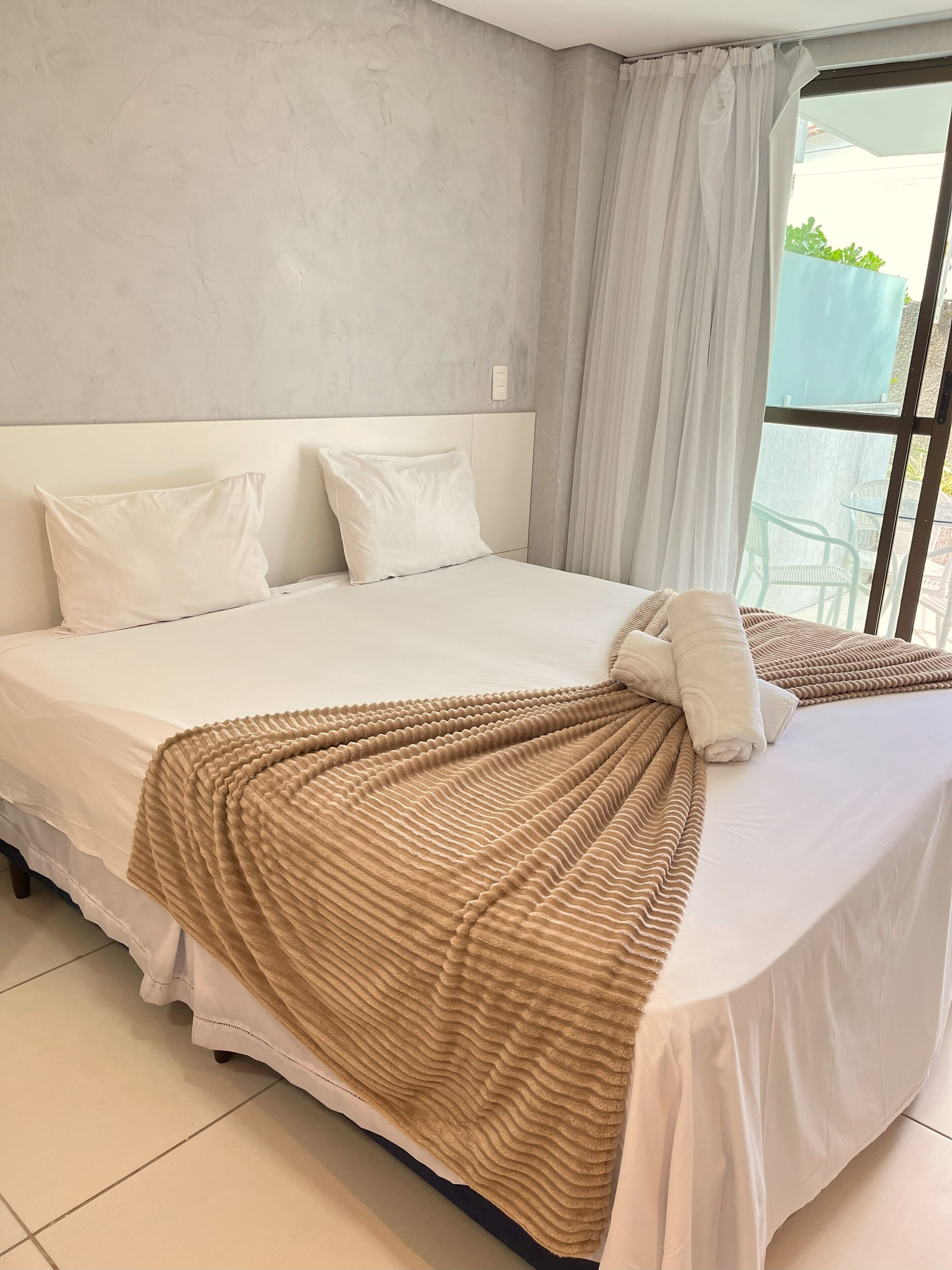 A neatly made bed with white linens and a textured brown blanket, a white headboard, and a window with a view of the outdoors.