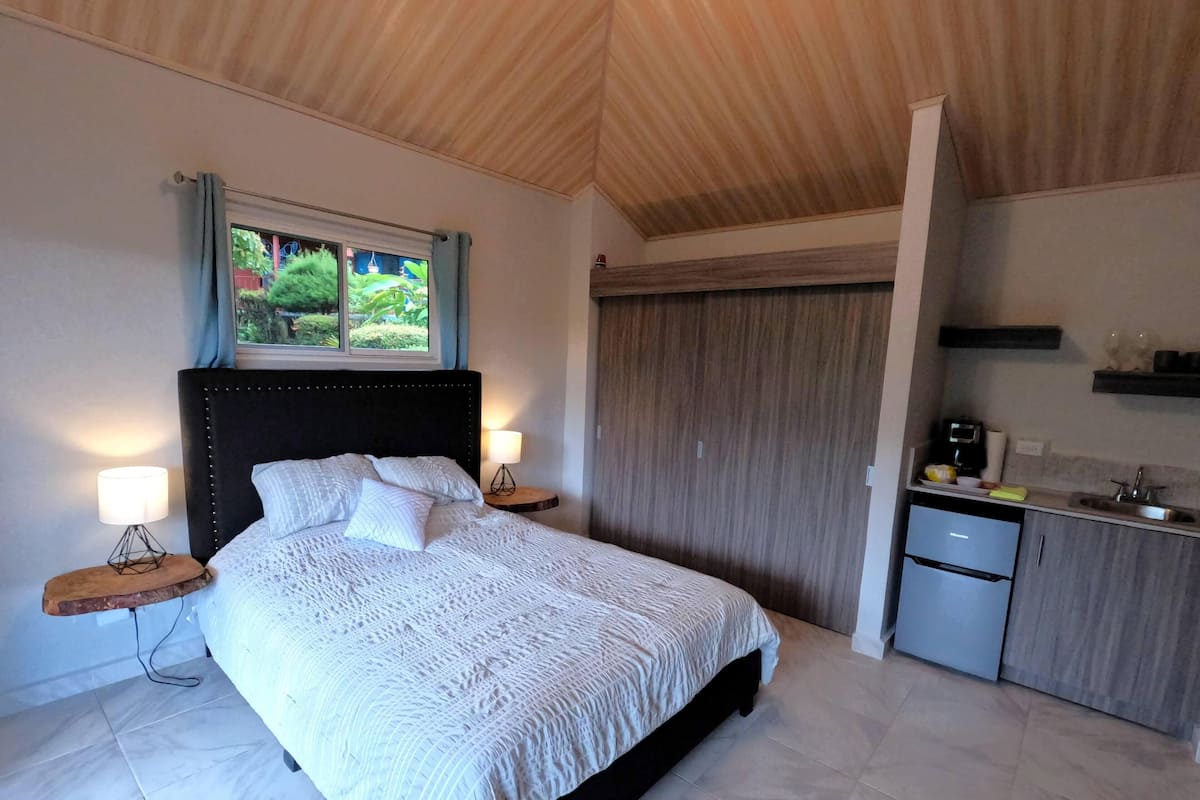 A bedroom with a large bed, bedside lamps, a window with a view of greenery, a wooden ceiling, and a small kitchenette.