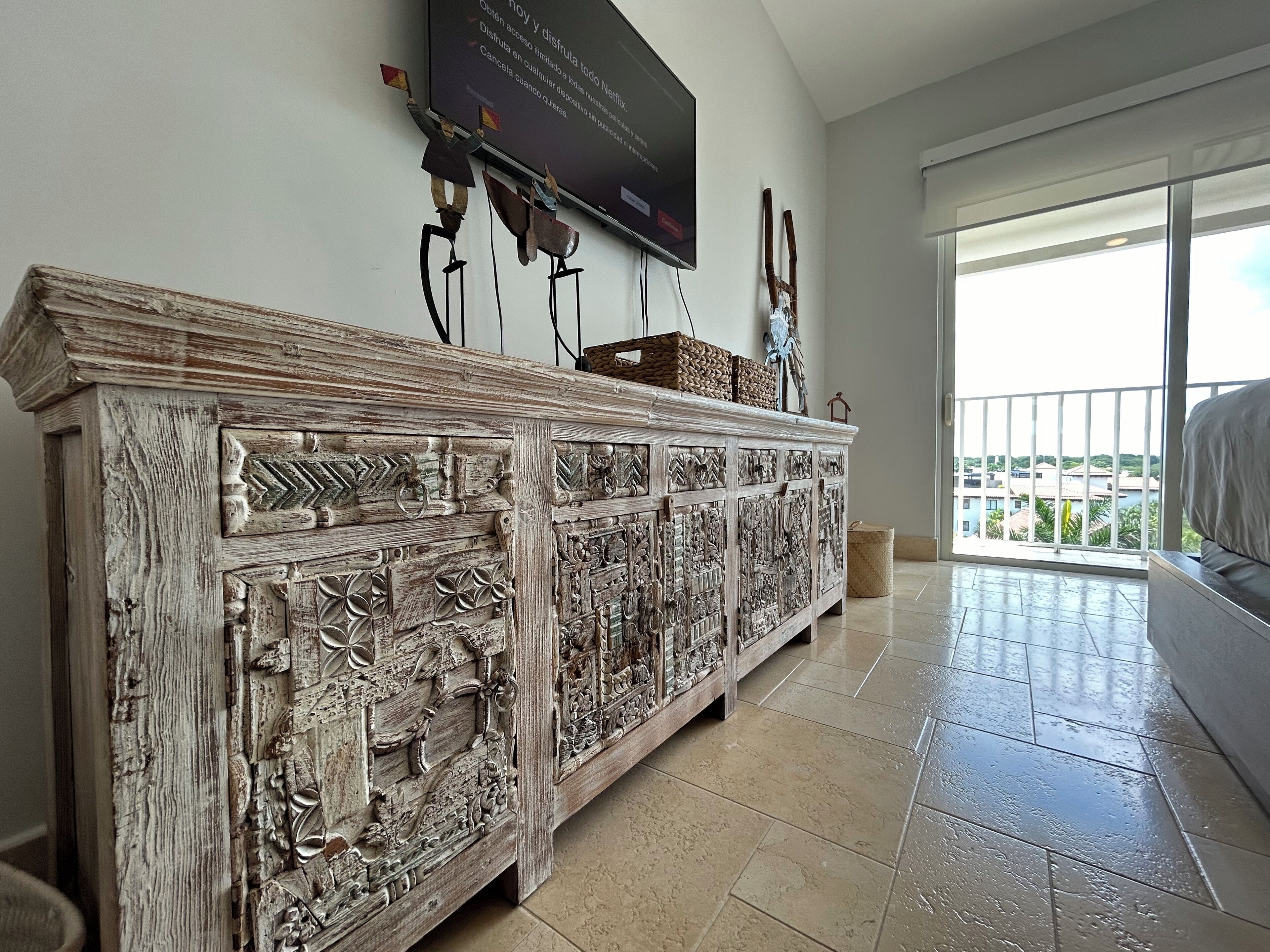 Uma sala de estar com um armário de madeira grande e ricamente detalhado, uma TV de tela plana na parede, e uma varanda com vista para edifícios e áreas verdes.
