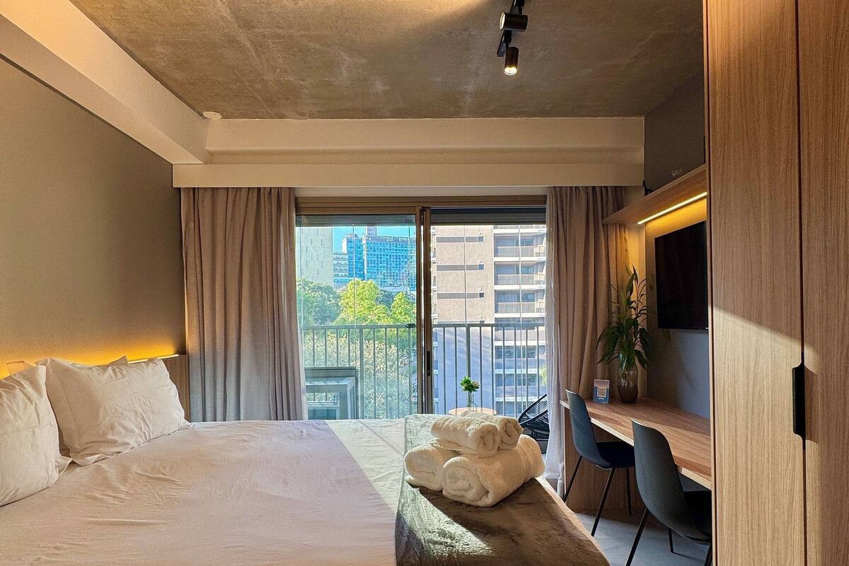 A modern hotel room with a large bed, a desk with a chair, a potted plant, and a view of the city through a balcony.