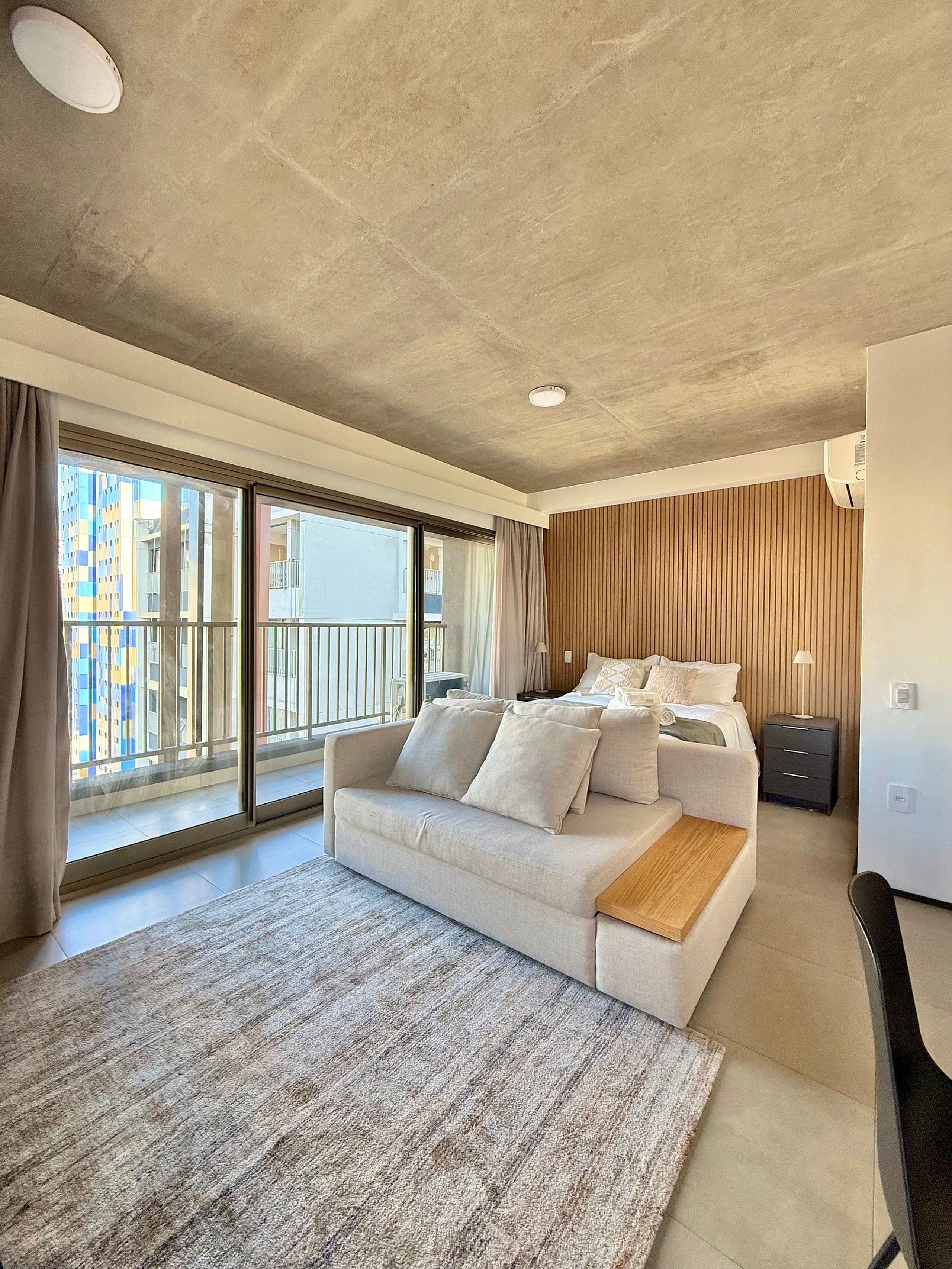 Ein modernes Hotelzimmer mit einem großen Bett, einer Couch, einem kleinen Schreibtisch und Blick auf eine Stadtlandschaft durch große Fenster.