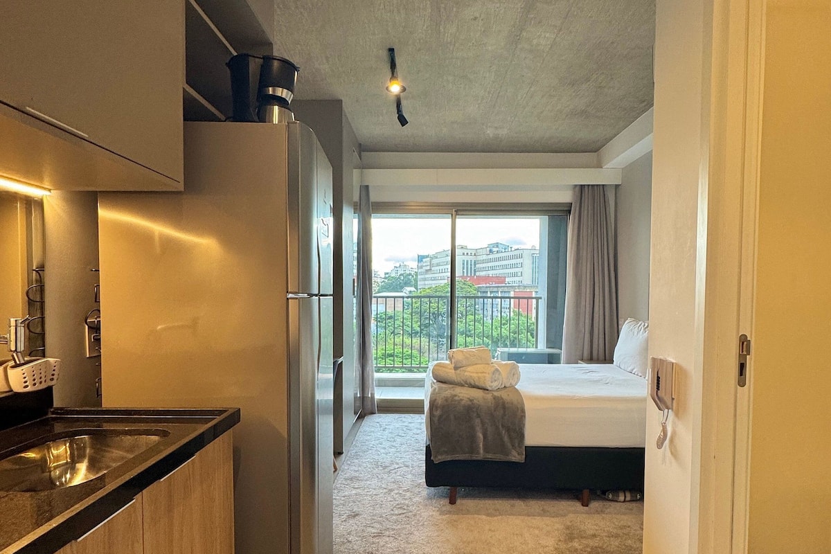 A modern hotel room with a kitchenette, a bed with a view, and a large window.