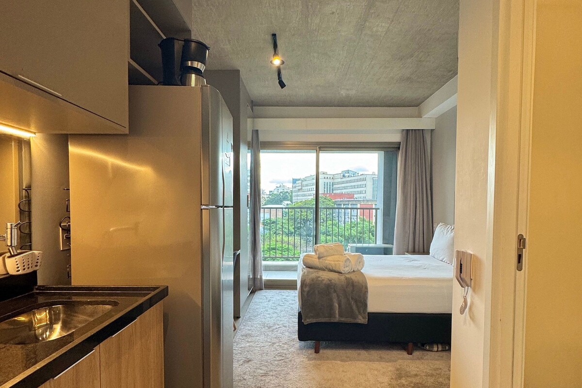 A modern hotel room with a kitchenette, a bed with a view, and a large window.