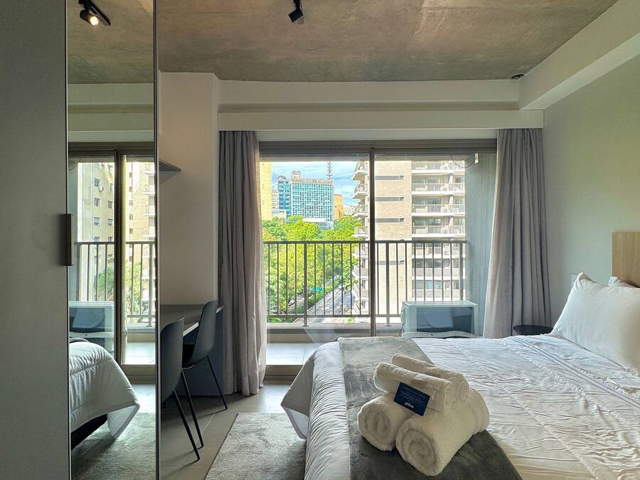 Habitación de hotel con cama, escritorio, silla, ventana grande con vistas a edificios y balcón.