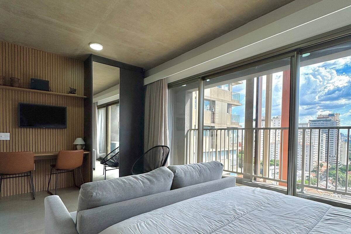 A hotel room with a large bed, a desk, a chair, a TV, and a balcony with a city view.