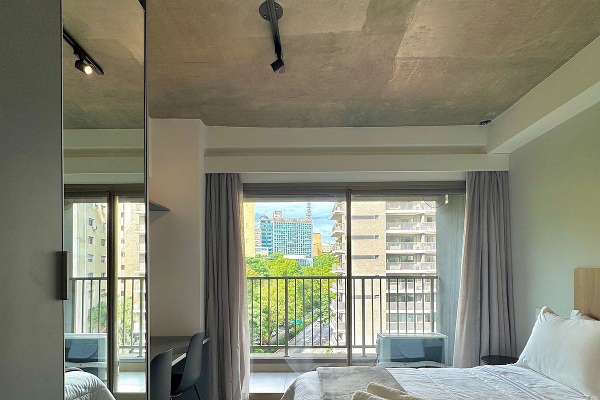 A hotel room with a bed, a desk, a chair, a large window with a view of buildings, and a balcony.