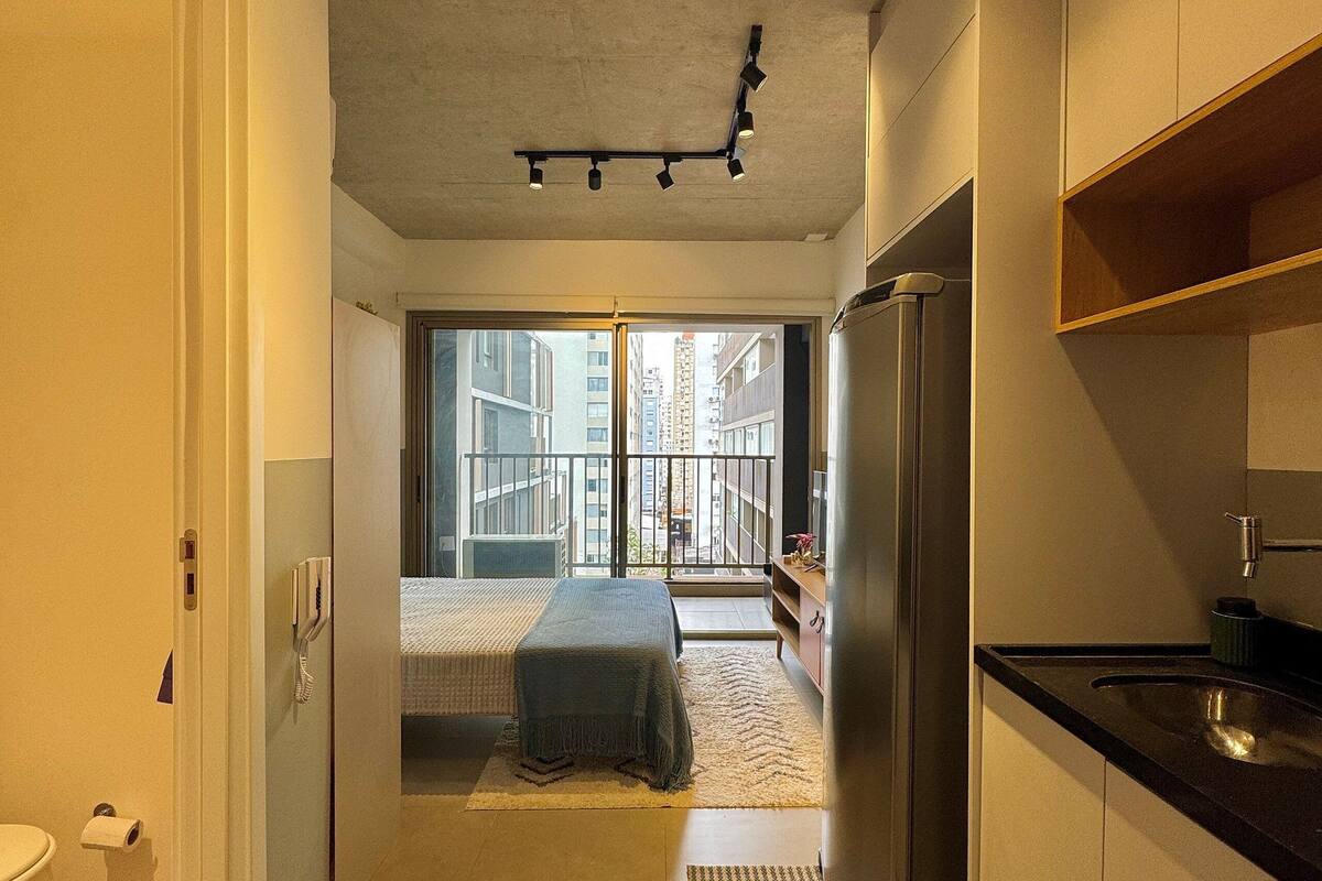 A narrow hallway leading to a bedroom with a large window, a kitchen area with a refrigerator, and a bathroom with a toilet.
