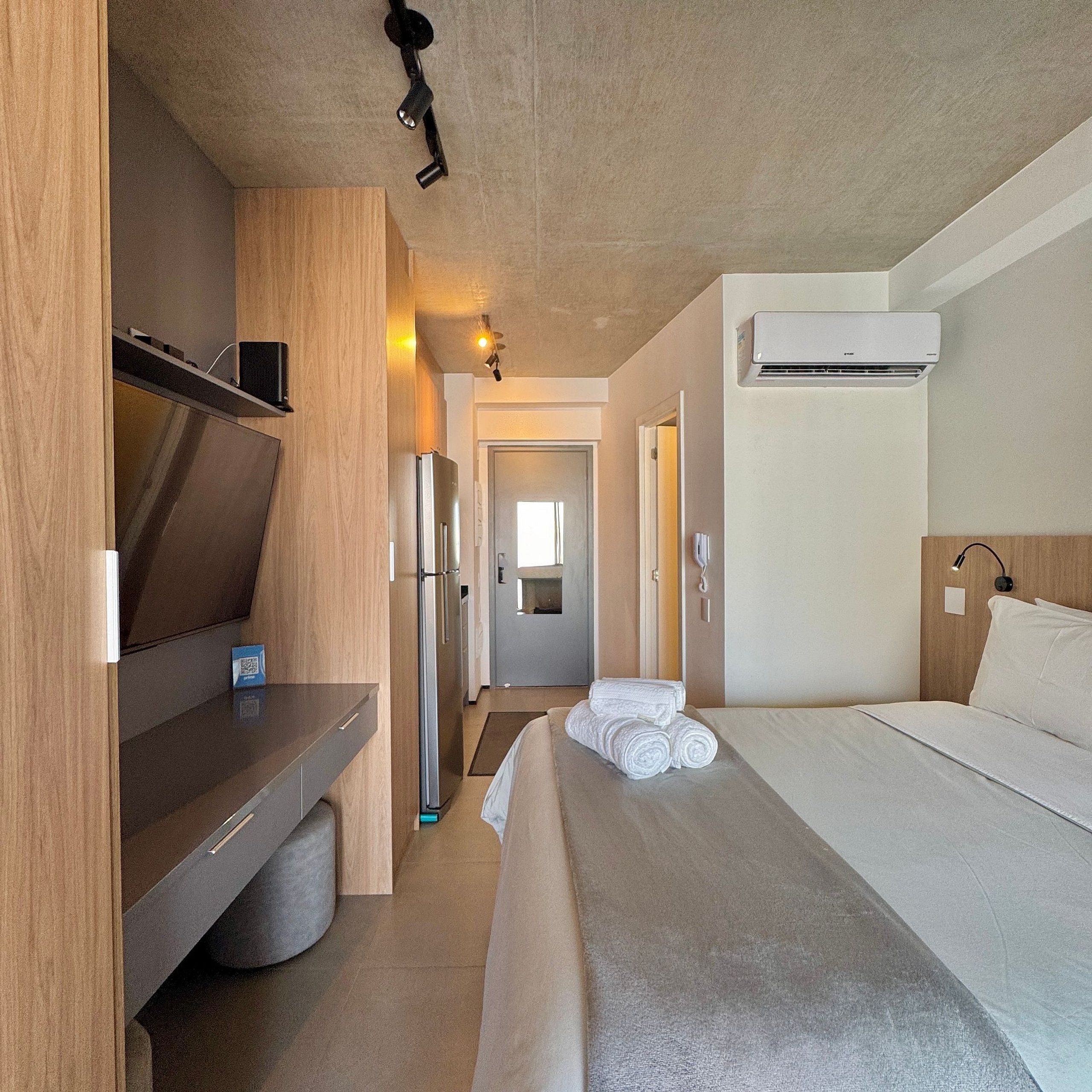 Quarto de hotel compacto com cama, pequena cozinha, frigorífico, micro-ondas, televisão de ecrã plano e unidade de ar condicionado.