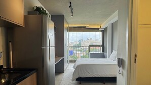 Habitación de hotel compacta con kitchenette, cama, teléfono junto a la cama y balcón con vista a la ciudad.