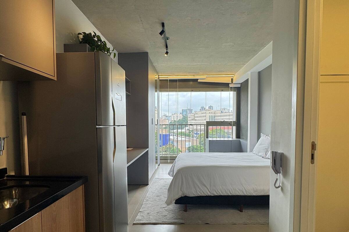 A compact hotel room with a kitchenette, a bed, a bedside phone, and a balcony with a city view.