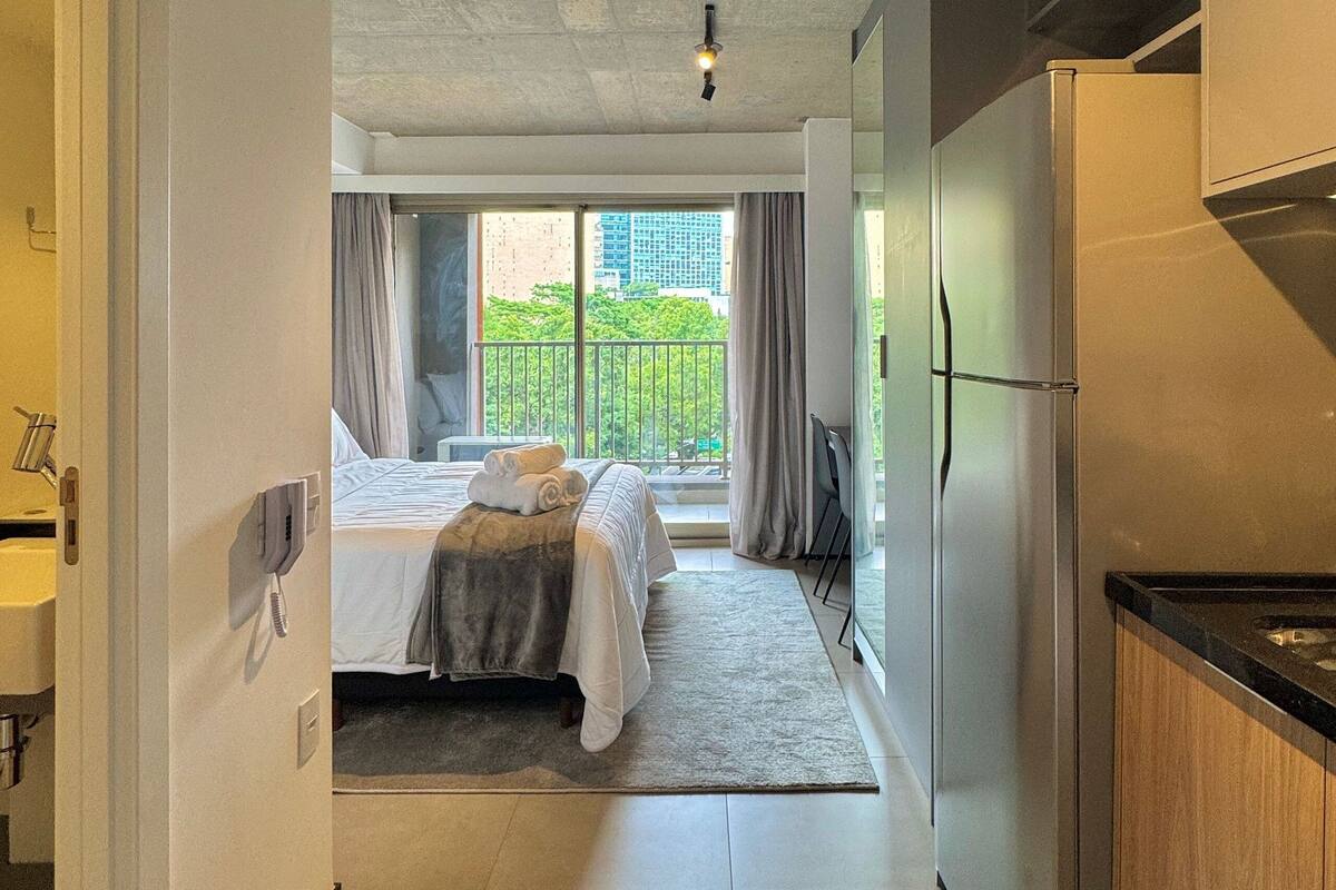 A hotel room with a bed, a desk, a refrigerator, and a view of a balcony with greenery.