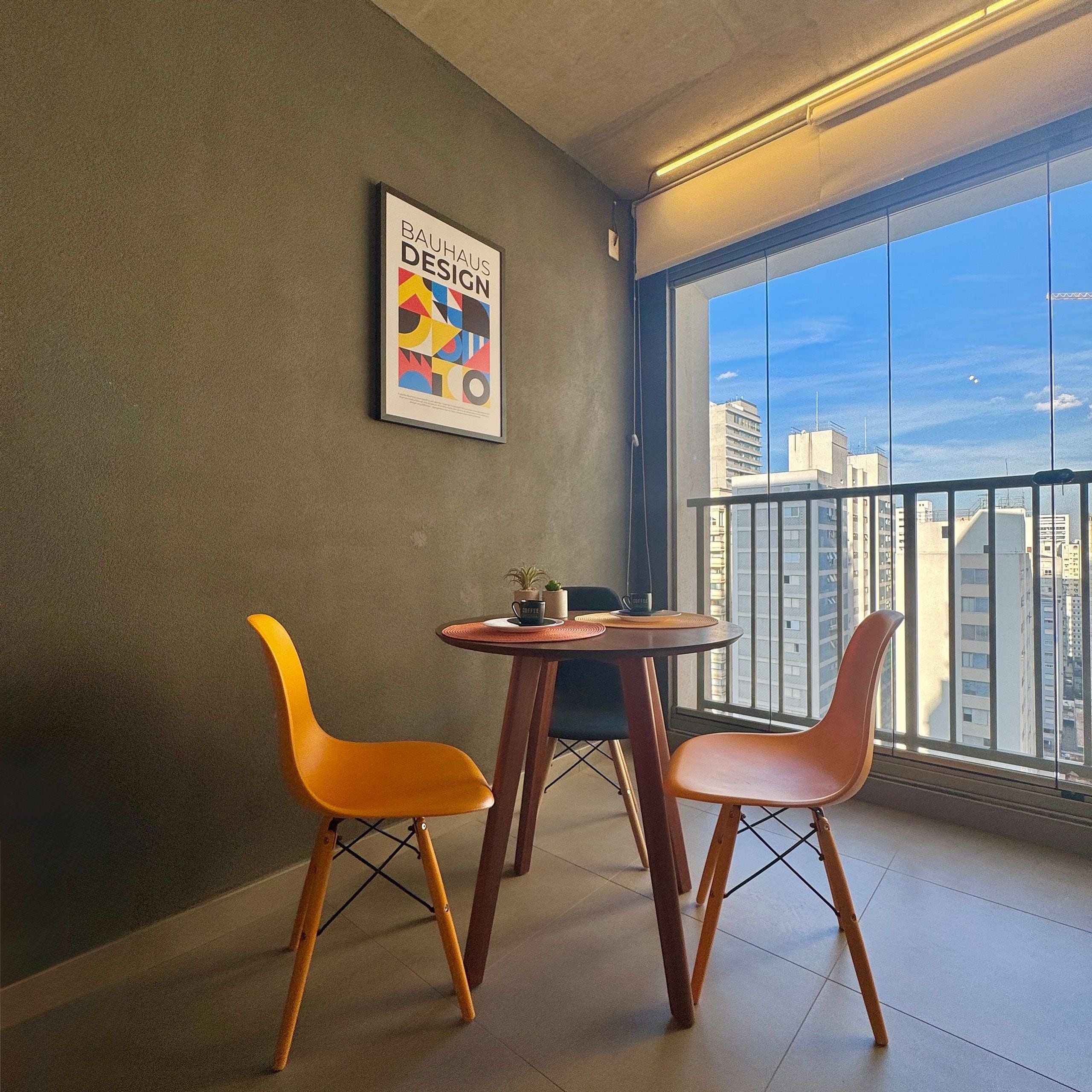 A modern room with a large window, two orange chairs, a small table with a coffee cup, and a Bauhaus Design poster on the wall.