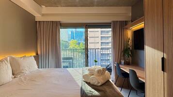 Quarto de hotel moderno com uma cama grande, uma escrivaninha com cadeira, uma planta em vaso e vista para a cidade através de uma varanda.