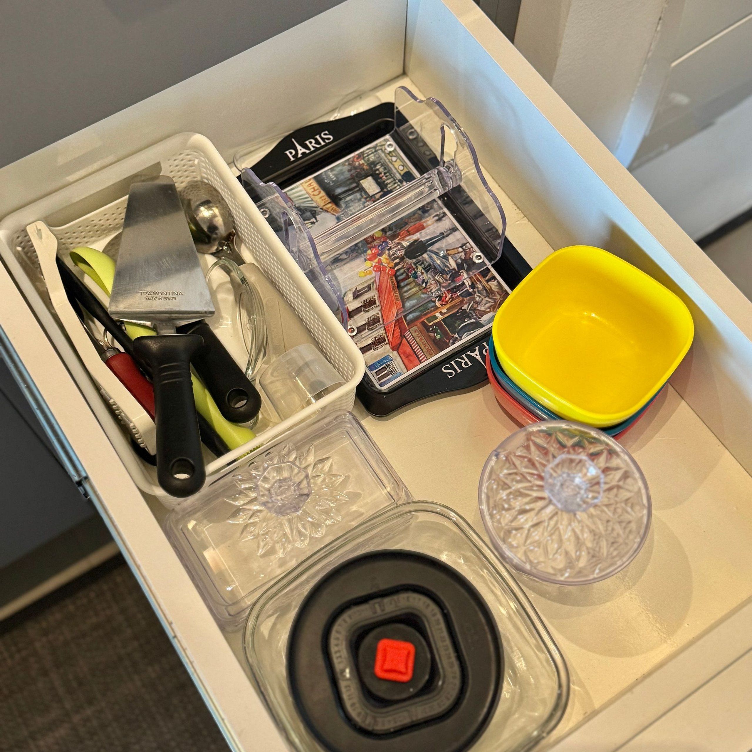 A drawer with kitchen utensils, a magazine, a yellow bowl, and a glass container.