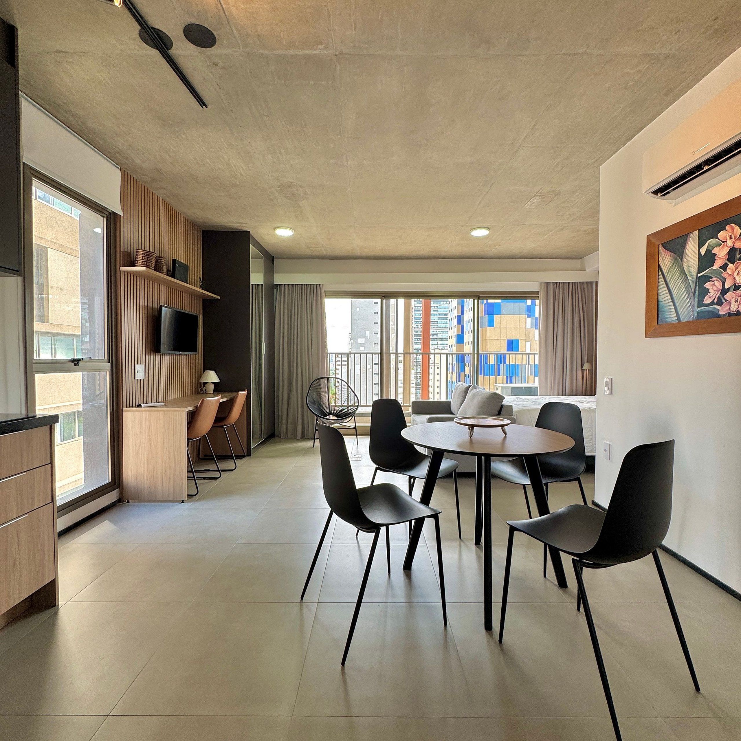 A modern hotel room with a dining area, a TV, a large window with a city view, and a painting on the wall.