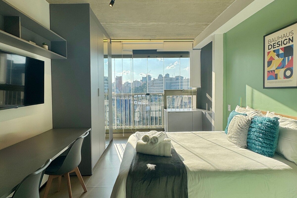 A modern bedroom with a bed, a desk, and a large window offering a city view.