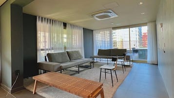 Un salon moderne avec un canapé, une table basse et des fauteuils, une grande fenêtre avec des rideaux et une vue sur les bâtiments à l’extérieur.