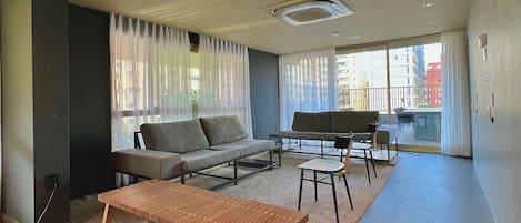 A modern living room with a sofa, coffee table, and chairs, a large window with curtains, and a view of buildings outside.
