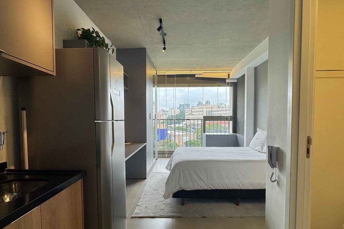 A compact hotel room with a kitchenette, a bed, a bedside phone, and a balcony with a city view.