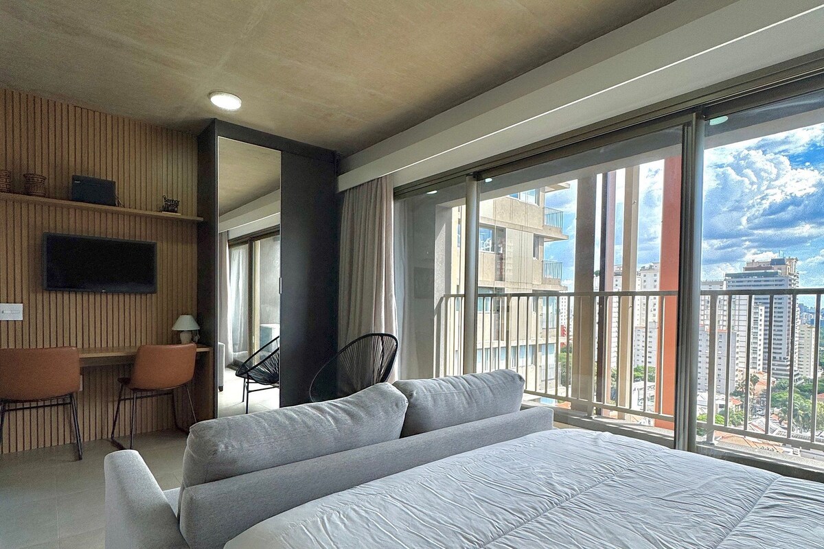 A hotel room with a large bed, a desk, a chair, a TV, and a balcony with a city view.
