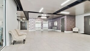 Eine geräumige Lobby mit weißem Sessel, einer Bank und einem Empfangstisch. Zwei Aufzüge und eine Chandelier sind vorhanden.