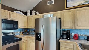A kitchen with light wooden cabinets, a stainless steel refrigerator, a microwave, and a stove. - Emily By The Sea by Ryson Vacation Rentals (Galveston)