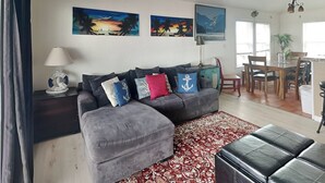 A living room with a grey sectional sofa, a red and white patterned rug, and a black leather ottoman. There are three paintings on the wall, a dining area with wooden furniture, and a chandelier. - Emily By The Sea by Ryson Vacation Rentals (Galveston)