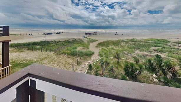 A beach view with a sandy path, a wooden fence, and a building in the foreground. - Emily By The Sea by Ryson Vacation Rentals (Galveston)