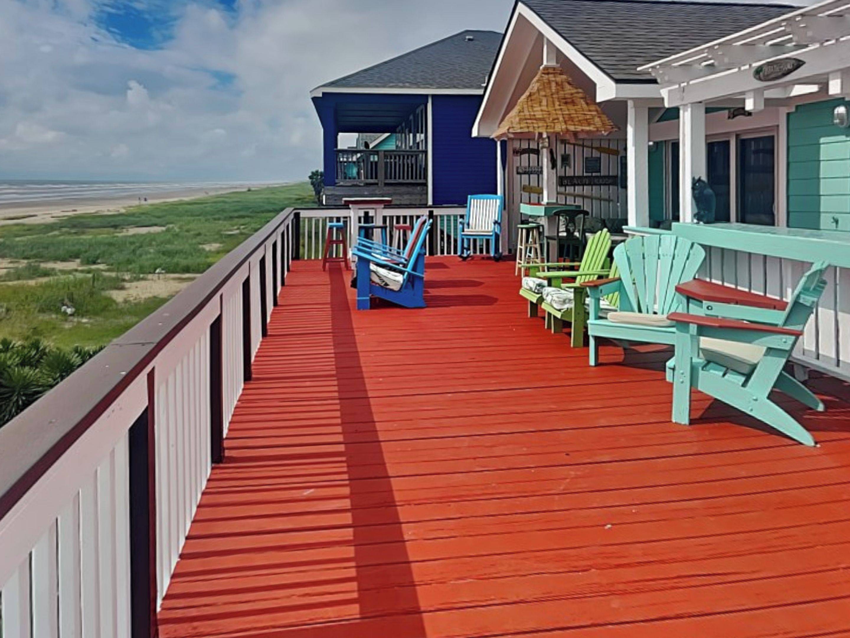 A wooden deck with red flooring, green and blue Adirondack chairs, and a blue building with a thatched roof.