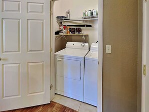 A laundry room with a washer and dryer, a shelf with various items, and a white door. - Alto Paradise by Ruidoso Vacation Rentals (Alto)
