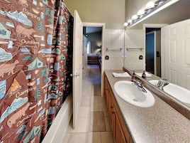 Un baño con cortina de ducha decorada con motivos de naturaleza y campamento, un lavabo doble y un espejo.