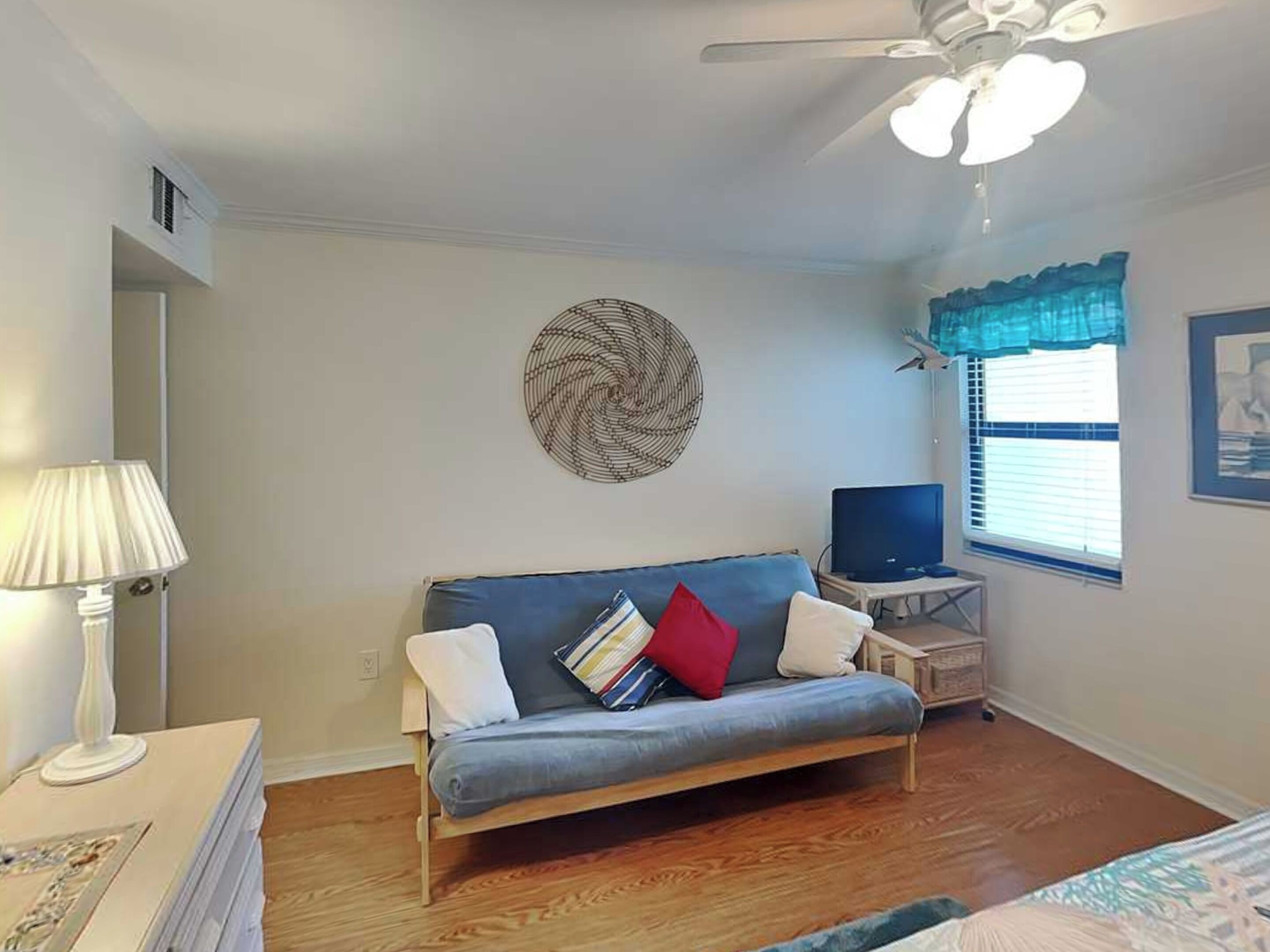 A room with a sofa, a television, a ceiling fan, a window with blinds, and a wall decoration.