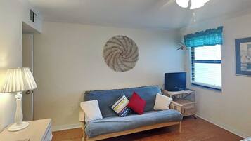 A room with a sofa, a television, a ceiling fan, a window with blinds, and a wall decoration.